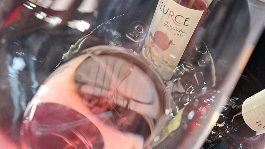 Primer pla d'una copa de vi rosat de color rosa pàl·lid, vista des d'un angle superior. Al fons, fora de focus, es veuen ampolles de vi rosat Ilurce de Garnatxa en una galleda de gel. L'etiqueta de l'ampolla central mostra el nom 'ILURCE'