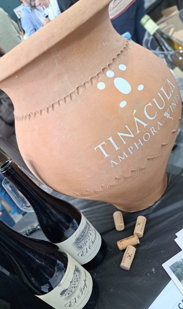 Primer pla detallat d'una autèntica tinaja de fang utilitzada per a l'elaboració artesanal de vi. S'hi llegeix la inscripció 'TINÁCULA AMPHORA WINES'. A la base, hi ha dues ampolles de vi 'El Imperio' amb etiquetes elegants que mostren el paisatge de vinyes velles, i suros sobre una estovalles fosca.