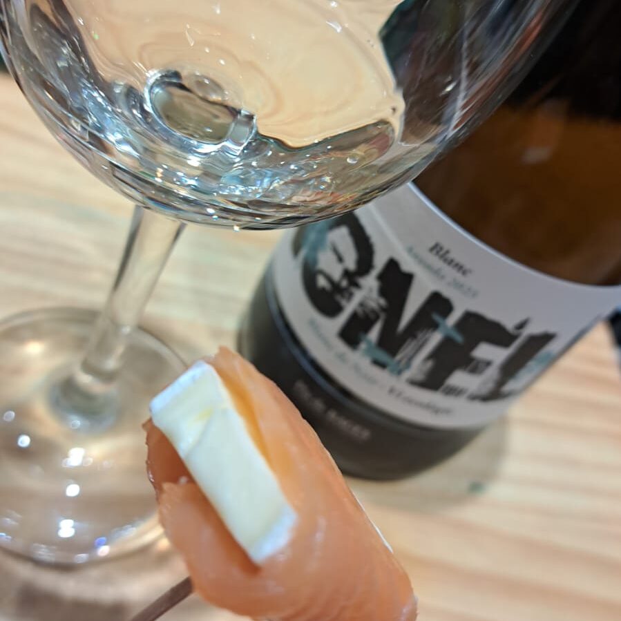 Un apetitós rollet de salmó amb formatge brie en primer pla, maridat al fons amb una copa i una ampolla del vi blanc de noirs Nonell de Vinalium Manresa i Celler Collbaix.