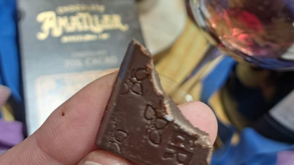 Una rajola de xocolata negra amb una mossegada en primer pla, acompanyada al fons d'una copa de vi Nonell i una elegant caixa de disseny modernista a Vinalium Manresa.